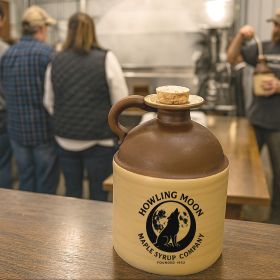 A Howling Moon Maple Syrup jug sits on a wooden counter inside a maple syrup factory with workers and equipment visible in the blurred background.