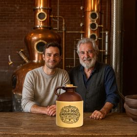 Two men, possibly distillers, sit at a wooden table inside a copper still distillery with an Old Dog Moonshine jug displayed prominently in front of them.Multiple Old Dog Moonshine ceramic jugs arranged neatly on a rustic wooden shelf in a warm, decorative shop environment.