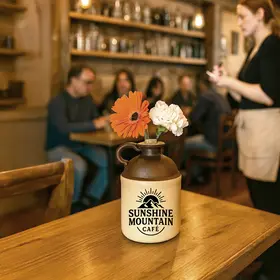 A ceramic moonshine jug labeled “Sunshine Mountain Café” sits on a wooden table being used as a flower vase with orange and white blooms, inside a cozy café where a server takes orders and customers sit at nearby tables.