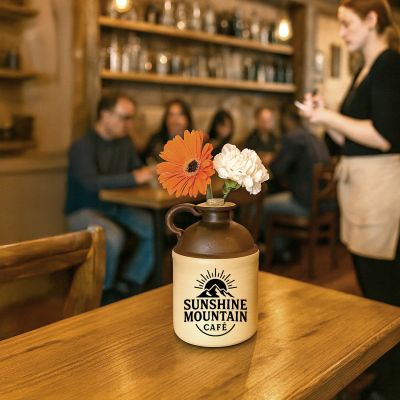 A ceramic moonshine jug labeled “Sunshine Mountain Café” sits on a wooden table being used as a flower vase with orange and white blooms, inside a cozy café where a server takes orders and customers sit at nearby tables.